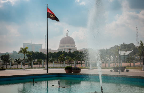 A fountain splashes water in the foreground while the Angolan flag flutters in the wind showcasing the presidential palace in Luanda under a clear sky.
